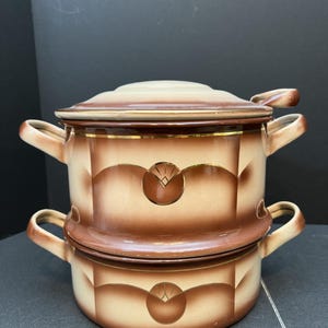 May include: Two-piece ceramic cookware set in shades of brown and beige. The set includes a lidded pot and a smaller pot stacked underneath. Each piece has a decorative design and gold trim.