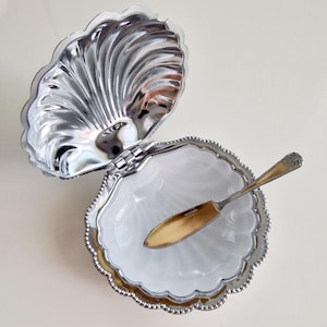 Könnte beinhalten: Eine silberfarbene Metallschale in Muschelform mit Klappdeckel und weißem Innenraum. Die Schale hat einen gewellten Rand und eine reflektierende Außenseite. Ein kleiner, passender silberfarbener Metalllöffel liegt in der geöffneten Muschel.