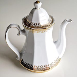 Może przedstawiać: Biały porcelanowy imbryk o sześciokątnym korpusie, zakrzywionym uchwycie i pasującej pokrywce. Imbryk posiada złote wzory kwiatowe i koronkowe wzdłuż podstawy, krawędzi i pokrywki, z pozłacanym uchwytem na szczycie pokrywki.