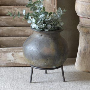 Op de afbeelding: Een ronde, donkergekleurde, getextureerde pot met een wijde opening, rustend op een dunne, driepotige zwarte metalen standaard. De pot bevat een takje met groene eucalyptusbladeren en staat naast een hoge, cilindrische, verweerde houten zuil.