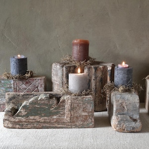 Op de afbeelding: Een verzameling brandende pilaar kaarsen in verschillende kleuren, waaronder grijs, bruin en wit, worden getoond op verweerde, gesneden houten blokken. De blokken zijn versierd met gedroogd mos en hebben een rustieke, antieke uitstraling. De achtergrond is een gedempt groen.
