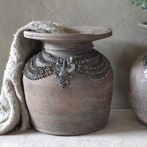 May include: Two large, rounded brown clay pots with wide rims. The left pot is draped with a textured beige fabric and adorned with a dark silver-toned multi-layered chain necklace. The right pot features a silver-toned circular link chain.