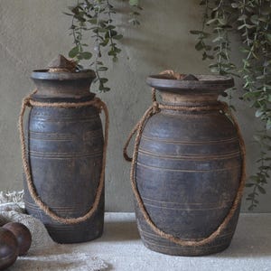 May include: Two dark brown wooden vessels with cylindrical bodies, flared rims, and thick jute rope handles attached to the necks. The containers feature horizontal carved grooves along the sides and are accompanied by four smooth, round wooden spheres.