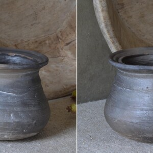 Może przedstawiać: Ciemnoszary, zaokrąglony garnek ceramiczny z szerokim, rozszerzającym się brzegiem i teksturowaną, matową powierzchnią. Naczynie posiada subtelne poziome żłobienia i r&oacute;żnice w odcieniach na korpusie, z głębokim, otwartym wlotem i stabilną podstawą.