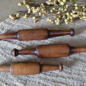 Può includere: Tre antichi mattarelli in legno con manici arrotondati, disposti su un tessuto grigio strutturato. I mattarelli sono di varie tonalità di marrone, con venature del legno visibili. Un rametto di verde con piccoli boccioli rotondi è sullo sfondo.