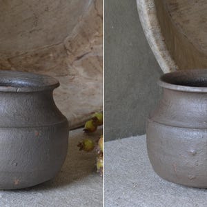 Może przedstawiać: Mały, ciemnobrązowy, zaokrąglony garnek ceramiczny z szerokim, rozszerzającym się brzegiem i teksturowaną, matową powierzchnią. Naczynie posiada wyraźne poziome żebrowanie wok&oacute;ł środkowej części oraz lekko zwężającą się podstawę o prostej formie.