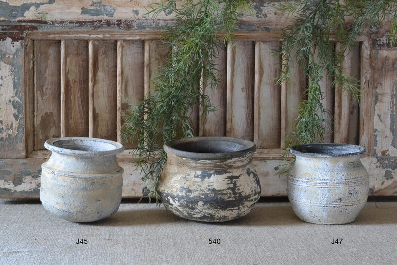 Może przedstawiać: Trzy okrągłe ceramiczne donice o teksturowanym, postarzanym wykończeniu. Lewa donica oznaczona jest jako J45, środkowa jako 540, a prawa jako J47. Każda donica posiada szeroki rant, zaokrąglony korpus oraz przetartą, wielotonową powierzchnię zewnętrzną.