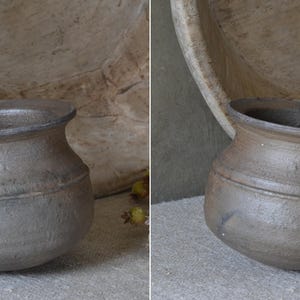 Może przedstawiać: Mały, ciemnoszary ceramiczny garnek o zaokrąglonym korpusie, szerokim otworze i rozszerzanym rancie. Powierzchnia ma teksturowane, matowe wykończenie z subtelnymi r&oacute;żnicami w odcieniach. Garnek posiada poziomą linię wok&oacute;ł środkowej części.