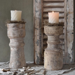 Pode incluir: Dois castiçais rústicos de madeira com velas brancas acesas. Os castiçais são feitos de madeira envelhecida e têm um acabamento desgastado. As velas são colocadas sobre uma cama de musgo seco. O fundo inclui uma veneziana de madeira desgastada.