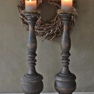 Può includere: Due alti candelabri in legno marrone scuro con un design tornito a fuso e una base ampia e arrotondata. Ogni candelabro sostiene una spessa candela a colonna grigio chiaro sormontata da un po' di muschio secco, davanti a una ghirlanda di rametti.