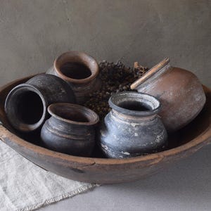 Peut inclure: Un grand bol en bois brun foncé contenant quatre petits pots en argile patinée dans des tons de noir, gris et terre cuite. Les pots présentent des textures variées et des formes arrondies, regroupés avec un amas de petits éléments botaniques séchés et sombres à l'intérieur du bol.