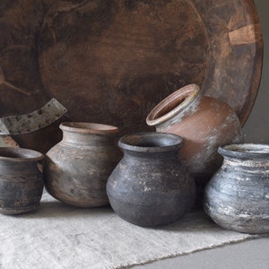 Könnte beinhalten: Eine Sammlung rustikaler, handgefertigter Töpferwaren in verschiedenen Größen und Grautönen, Braun und Schwarz. Die Töpferwaren haben ein verwittertes, gealtertes Aussehen, das auf eine Vintage- oder Antik-Ästhetik hindeutet. Im Hintergrund befindet sich ein großer, runder Holzkorb.