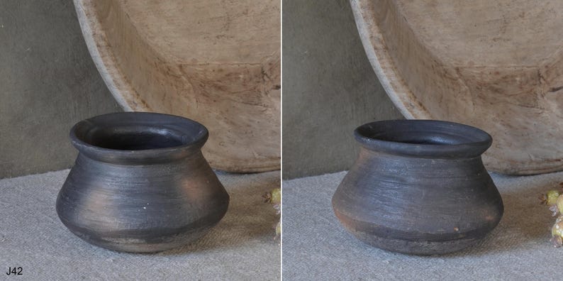 Może przedstawiać: Mały, ciemny, antracytowy garnek ceramiczny o szerokim, zaokrąglonym korpusie i rozszerzanym rancie. Powierzchnia ma matowe, teksturowane wykończenie z subtelnymi poziomymi prążkami. Tekst J42 jest nadrukowany w lewym dolnym rogu obrazu.