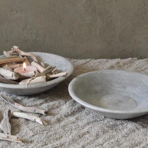 Può includere: Due ciotole di pietra grigia, una riempita di legno alla deriva e una candela accesa. L'altra ciotola è vuota. Le ciotole sono su una superficie strutturata dai toni neutri, suggerendo un'estetica naturale e minimalista. Ideale per l'arredamento della casa.