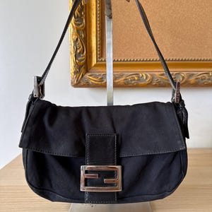 May include: Black shoulder bag with a silver buckle. The bag features a flap closure and a single shoulder strap. The bag is made of a dark fabric and has a rectangular shape.