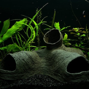 May include: A dark grey ceramic cave-like structure with three openings, surrounded by green aquatic plants in a black gravel tank.