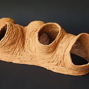 Può includere: Una scultura in terracotta con una superficie testurizzata e tre grandi aperture. La scultura è di colore marrone chiaro e ha una forma organica unica.