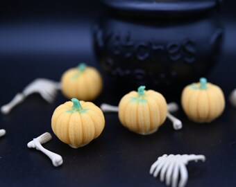 May include: Four small yellow pumpkin-shaped sweets with green stems, surrounded by white plastic skeleton bones on a black surface. A black cauldron is in the background.