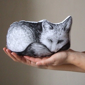 May include: A black and white plush pillow shaped like a fox curled up sleeping. The fox has a white muzzle and a black nose.