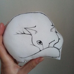 May include: A small, white, rounded plush toy featuring a black line drawing of a curled-up fox. The fox is depicted in a sleeping position with its tail wrapped around its body, showcasing simple, clean ink-style outlines on a soft fabric surface.