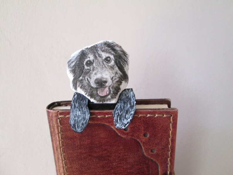 May include: A handmade dog bookmark featuring a smiling dog illustration. The dog's head and paws are visible, appearing to peek over the top of a brown leather-bound book. The bookmark is designed to be inserted into a book.