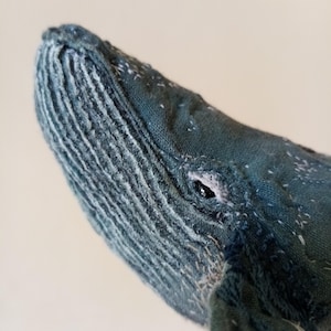 Puede incluir: Primer plano de un peluche de ballena azul hecho a mano. La cabeza de la ballena está detallada con tela texturizada, costuras visibles y un ojo pequeño y oscuro. El peluche tiene un aspecto suave y artesanal.