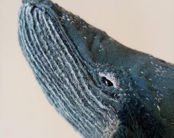 May include: Close-up of a handmade blue whale plush toy. The whale's head is detailed with textured fabric, visible stitching, and a small, dark eye. The plush has a soft, handcrafted appearance.