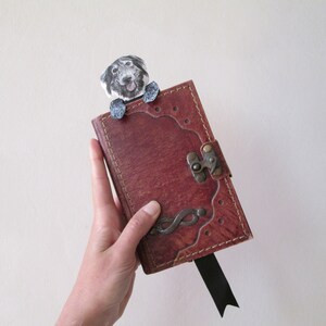 May include: A handmade leather journal with a decorative metal clasp and a black ribbon bookmark. A whimsical dog-shaped bookmark peeks over the top of the journal. The journal is a rich, reddish-brown color.