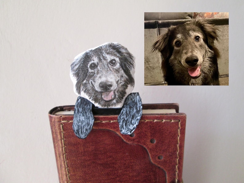 May include: A handmade dog bookmark featuring a smiling dog face and paws, appearing to peek over the top of a book. The bookmark is attached to a brown leather-bound book. A photo of the dog is in the upper right corner.