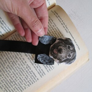 May include: A handmade bookmark featuring a watercolor illustration of a puppy. The black ribbon bookmark is placed between the pages of a book. The puppy has a black and gray coat and a pink tongue.