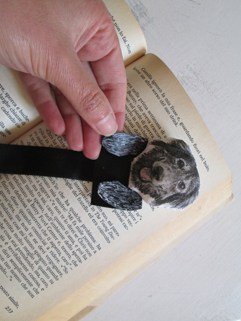 May include: A handmade bookmark featuring a watercolor illustration of a puppy. The black ribbon bookmark is placed between the pages of a book. The puppy has a black and gray coat and a pink tongue.