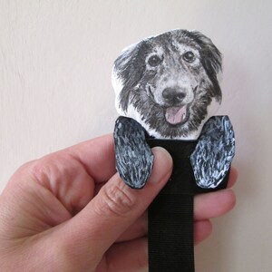 May include: A black and white dog-themed bookmark. The bookmark features a smiling dog face with a pink tongue and two black paws. The dog's face is printed on a white background, and the paws are painted black and white.