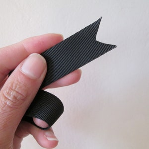 May include: A black ribbon with a notched end, held by a hand. The ribbon has a ribbed texture and is a solid, dark color. The ribbon's end is cut into a decorative shape.