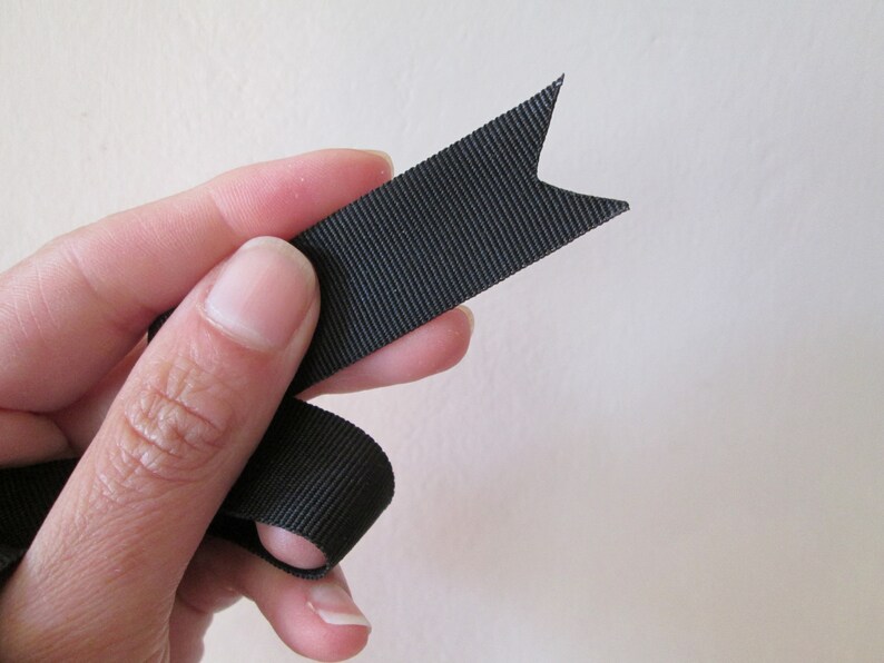 May include: A black ribbon with a notched end, held by a hand. The ribbon has a ribbed texture and is a solid, dark color. The ribbon's end is cut into a decorative shape.