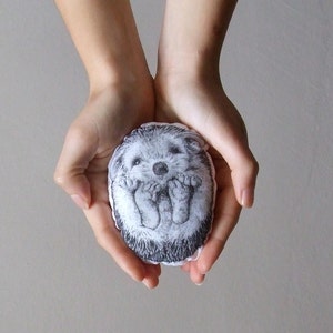 May include: A small, gray and white hedgehog-shaped stuffed animal with a black embroidered face and feet. The hedgehog is being held in the palms of two hands.