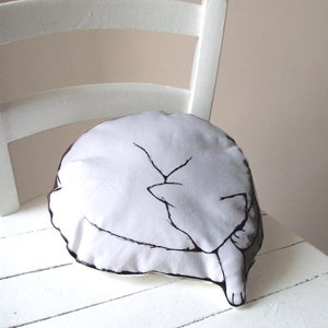 May include: A white pillow with a black outline of a cat curled up sleeping. The pillow is on a white wooden chair.