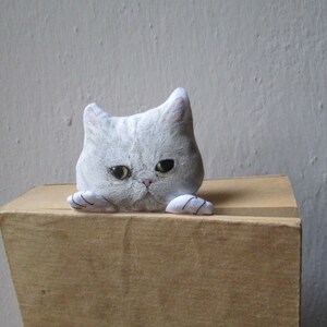 Peut inclure: Une figurine de chat en c&eacute;ramique blanche avec des yeux noirs et une expression renfrogn&eacute;e. Le chat regarde par-dessus le bord d'une bo&icirc;te en bois.