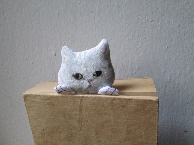 Peut inclure: Une figurine de chat en c&eacute;ramique blanche avec des yeux noirs et une expression renfrogn&eacute;e. Le chat regarde par-dessus le bord d'une bo&icirc;te en bois.