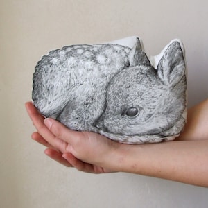 May include: A soft, plush pillow with a black and white illustration of a fawn sleeping. The pillow is held in a person's hands.