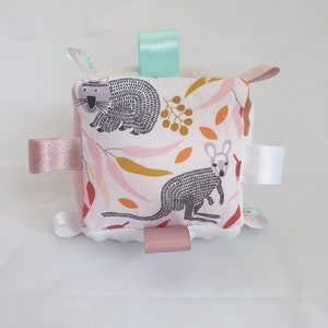 May include: A soft, white, square-shaped baby toy with a pink, white and brown fabric cover. The fabric features a pattern of leaves, berries, and two animals: a koala and a kangaroo. The toy has four fabric loops in pink, white and light blue.