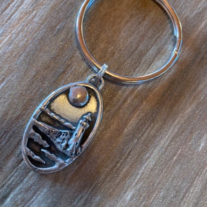 May include: Silver keyring with a detailed oval charm featuring a lighthouse on a cliff with a moon and clouds in the background.