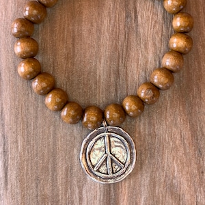 May include: A brown wooden bead bracelet with a silver peace sign charm. The bracelet is on a wooden background.