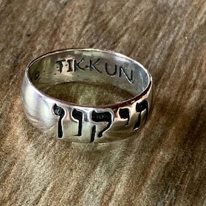 May include: A silver ring with the word "TIKKUN" engraved on the inside and Hebrew lettering on the outside.