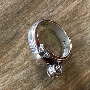 May include: A silver ring with a textured band and a decorative design on the side. The design features a heart shape with a small circle on top.