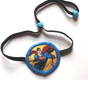 May include: A blue eye patch with an image of Superman flying. The eye patch is attached to a black elastic band with two blue plastic beads.