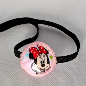May include: A circular fabric eye patch featuring a cartoon mouse with a red polka dot bow on a light pink background. The patch is attached to a black elastic strap with one red and one light pink plastic bead at the end of the band.