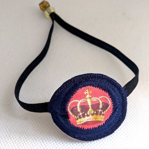 Op de afbeelding: Een blauw ooglapje met een elastisch bandje en een rode cirkel met een gouden kroonontwerp.