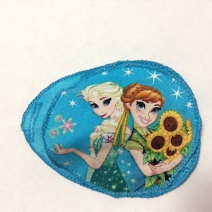 May include: A blue fabric patch featuring an image of two girls from the film Frozen. The girls are holding sunflowers. The patch has a white background with snowflakes.