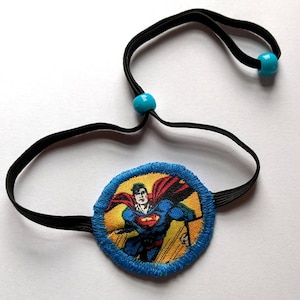 May include: A blue and yellow eye patch with a Superman image. The eye patch is attached to a black elastic strap with turquoise beads.