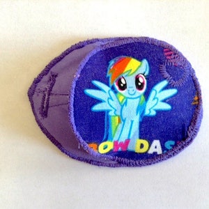 May include: Purple fabric eye patch with a Rainbow Dash pony appliqué. The patch features a blue pony with a rainbow mane and tail, and the text "Rainbow Dash" in yellow, red, and blue letters.